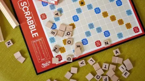 A Hasbro Scrabble board game arranged on a table alongside the box, tiles, and racks.