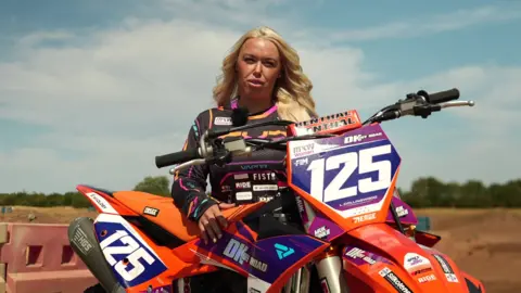 A woman with long blonde hair and wearing a long-sleeved black sports top with white logos stands behind a large orange motorbike, decorated with large white logos and branding. 