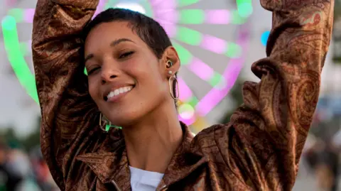 A young woman dances at dusk in front of a neon-lit Ferris wheel at a music festival. As well as large hoop earrings, she wears a rainbow-coloured ear-plug as protection for her hearing.