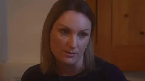 Alison Goldney seen sitting indoors with a wooden door visible in the background.