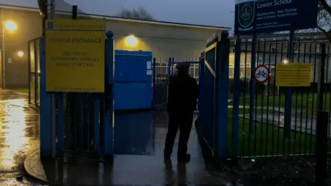 A police officer stands at the entrance gates of Kingsbury High School in Brent at dusk, with school signs visible and wet pavement reflecting light after rain.