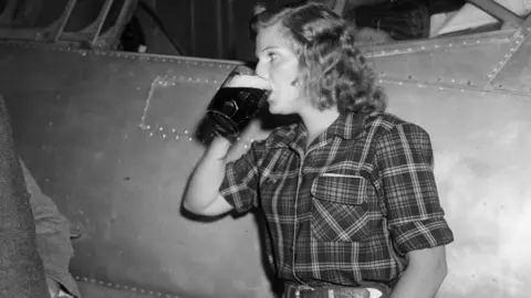 Getty Images A black and white photo of Richarda Morrow-Tait standing beside her aeroplane, wearing a plaid shirt and knocking back a pint