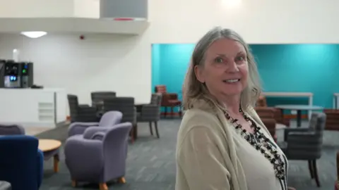 Volwarhampton Catherine appears to be the camera of the University of Lamond, which is standing in a room with white and blue walls. Chairs and tables are located behind him. She wears a light top and cardigan and deep beaded necklace.