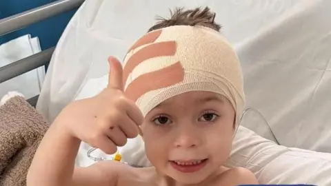 Courtney A young boy with a bandage around his head is in a hospital bed and has his thumbs up