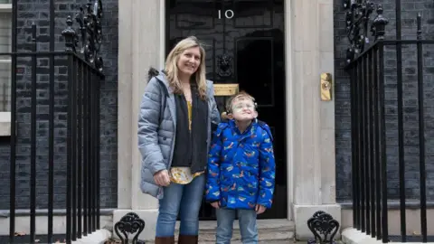 Claire Reece/Sense Claire Reece and Hugo outside the black shiny door of 10 Downing Street. Claire is on the left and wearing a grey puff coat over a blue scarf and jeans, her blonde hair on her shoulders. Hugo is wearing a bright blue jacket and jeans. 