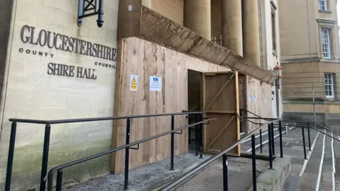 The outside of the historic Shire Hall. It is a large stone building, with steps and an access ramp in front of it. There is a large wooden hoarding covering the entrance, with warning signs on it. 