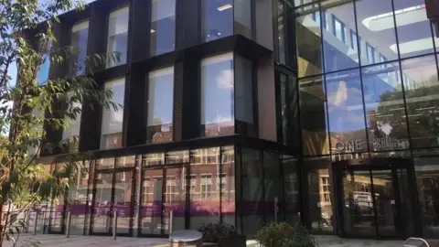 West Northamptonshire Council headquarters in Northampton. A large glass-panelled building with green plants in front of its door. 