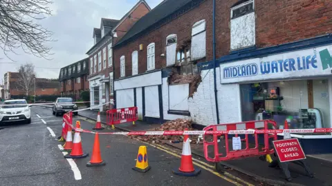 BBC A section of wall has come apart from the main structure with bricks covering a footpath. There is red tape and orange cones marking out the area and a red sign that reads "footpath closed ahead". 