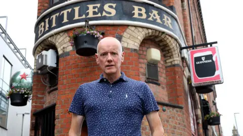 Pacemaker A bald man wearing a navy patterned polo shirt stands in front of a red bricked building with a black sign saying Bittles Bar in gold letters. There are Guinness and Heineken signs hanging off the sides of the narrow building.