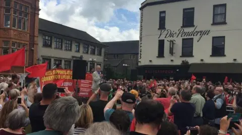 AFP Jeremy Corbyn speaks to a crowd of supporters at a rally at Merthyr Tydfil's Penderyn Square. He is standing at a podium and wearing a light suit and light blue shirt. Prominent in the background to the right of the picture is a large pub with a cream wall with the Welsh words Y Dic Penderyn in black paint. 