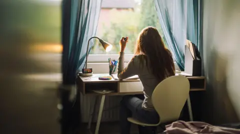 Getty Images A teenage girl doing homework while sitting at desk in home. She is sat on a white chair and the lamp on the desk is on. There is a window nearby.
