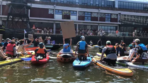 Surfers Against Sewage A group of people on paddleboards taken from the back. They are in a harbour holding signs to a crowd of people on the harbour side. They have multicoloured boards and life jackets.