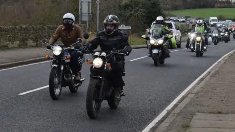 Heather Burton Two bikers lead a procession of motorcycles along a road. Other vehicles are visible in the line.