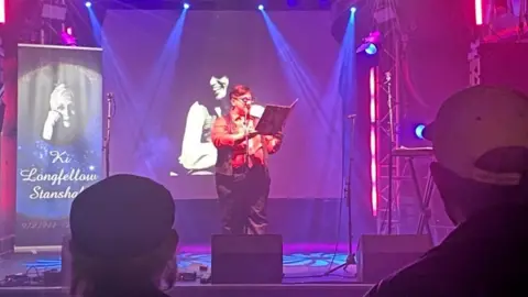 Sydney Longfellow Sydney standing onstage with a banner showing her mother to the side of the stage