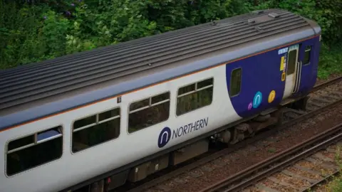 A Northern Trains carriage with its white and blue colours on a railway track with thick trees behind.