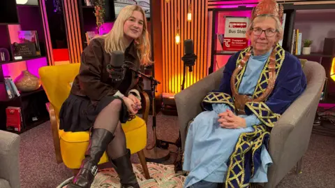 Danni is sitting in the podcast studio with the Grand Bard of Cornwall Jenefer Lowe. They are both smiling. Danni is wearing black biker boots, a black mini-skirt and brown cropped jacket. Jenefer is wearing a pale blue floor length gown under a blue velvet robe with gold brocade. She has a type of leather hat and a thick leather strap hanging around her neck. The studio is brightly coloured and lit with books, plants, vases and a bus stop sign on shelves. 