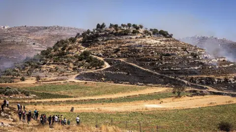 File photo showing Palestinians looking on as a fire burns on a hilltop that was seized by Israeli settlers near the town of Sinjil, in the occupied West Bank (4July 2025)