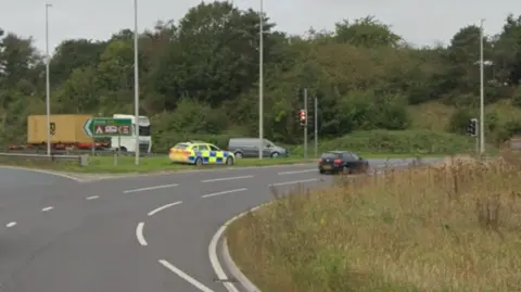 A Google Maps street view image of the Dock Spur roundabout in Felixstowe. A police car is parked up on the left and a black car is parked at the traffic lights. In the distance is a grey van and a large white and yellow lorry.