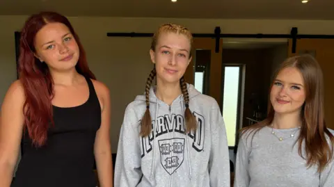 Luke Deal/BBC Three teenage girls smile at the camera. The girl on the left has long red hair and wears a black vest top. The girl in the middle has braided blonde hair and wears a grey hoodie. The girl on the left has long brown hair and wears a grey long sleeved top. 