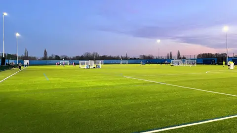 A 3G artificial football pitch, pictured at sunset, with floodlights illuminating it. There are players and goalposts dotted around the pitch, and trees in the distance at the edge of the pitch area.