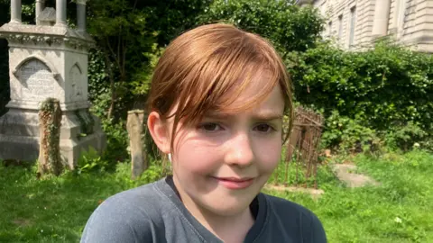 A young child with red hair and a grey t-shirt is in an old graveyard