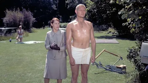 Getty Images Robert Shaw has short, dyed blonde hair and is dressed only in a small towel. He is standing in a garden surrounded by exercise equipment and is being admired by a woman with red hair and a grey suit. She is wearing sinister black gloves.