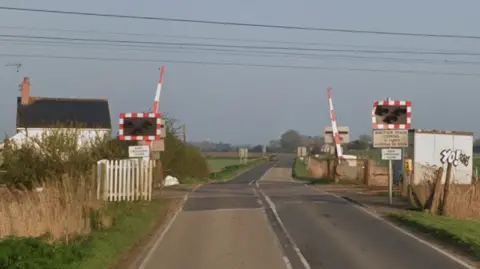 Google A road goes through a level crossing, which is in the middle of fields. There is a small white house on the left and a small hut on the right. There are lights and a barriers, which are open.