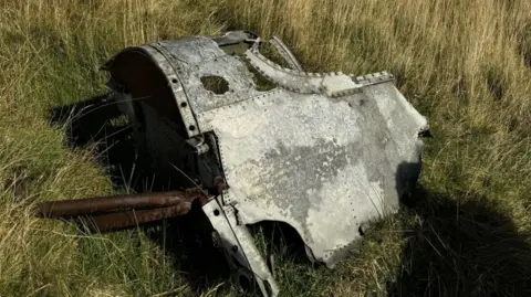 Jonny McNee A rusty silver ands bronze wreckage of a cockpit lying on green grass