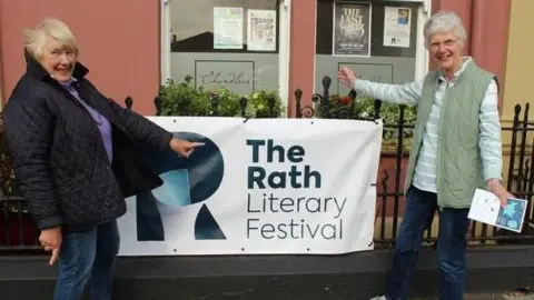two white-haired women gesturing to display a banner that reads Rath Literary Festival. They are smiling and looking at the camera