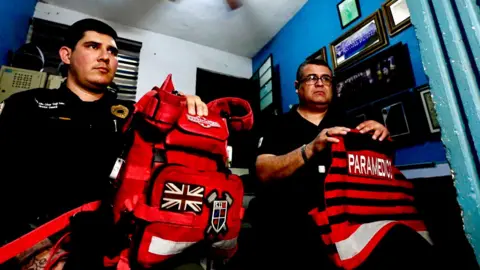 Darren Conway/BBC Two paramedics, Julio César Vega and Héctor Torres, sit in a small room. They are holding their kit on their knees. Both are wearing black uniforms and Torres is wearing glasses. 