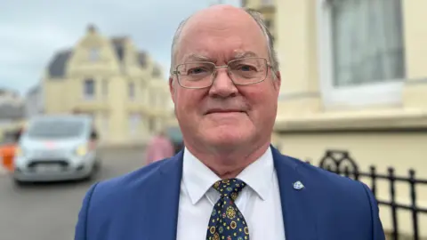 Simon Crowcroft - a man wearing silver rectangle framed glasses, a navy suit with a white shirt underneath and a navy tie with yellow, light blue and red flower looking symbols on it