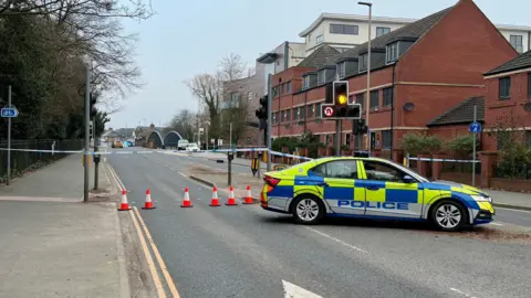 View looking down Abbey Park Road, with a police car parked across the highway in front of police tape and cones.