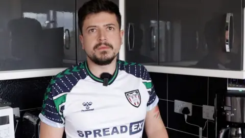 Benn Goodings standing in is kitchen wearing a white sports top. He has dark hair and a dark goatee-style beard.