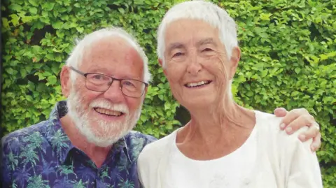 Malcolm Ridge Malcolm Ridge stood on the left grinning, he has white hair and a white beard and mustache. He is wearing black framed glassed and a purple and green palm tree patterned shirt. He has his arm around Ruth who also has white hair and is smiling at the camera. She is wearing a white top. 