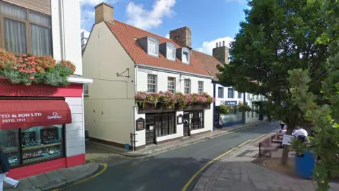 Google Street View image of Dumaresq Street in St Helier in Jersey with several shops on the left side of the road and trees and benches on the opposite pavement.