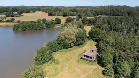 The Landmark Trust An aerial view of the control tower on the edge of a grassy, tree-fringed lawn which borders a lake with curved sides.