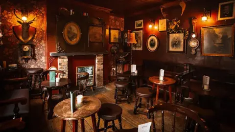 1936 Pubco The inside of Dickensian-style pub, The White Hart, which includes a stag's head mounted on the wall to the left of the fireplace. There is a number of small tables and wooden chairs and brown leather stools with the walls filled with paintings and ornaments.