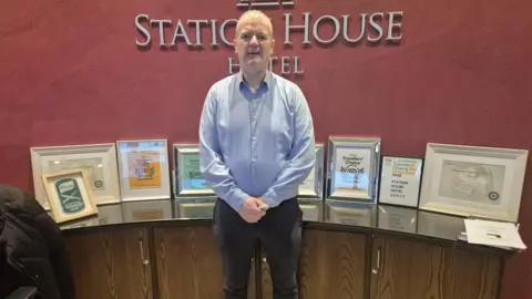 Brian Gallagher is wearing a blue shirt and black trousers. he is standing in The Station House Hotel reception. A number of framed pictures are near him. 