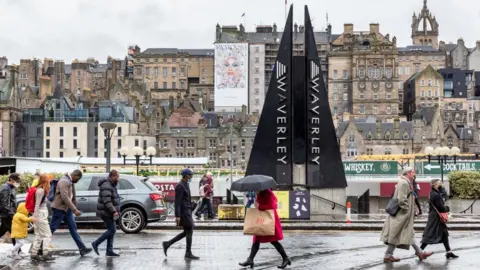 PA Media Commuters cross the street in front of the old town as showers and strong wind struck Edinburgh