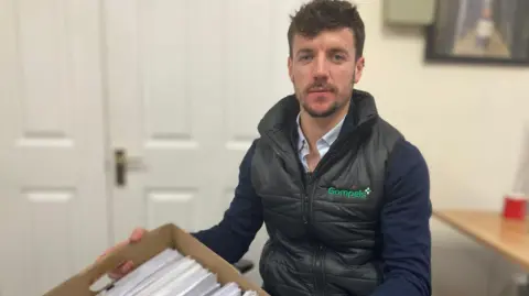 Mr Gompels looks at the camera wearing a black branded gilet and blue jumper holding a box