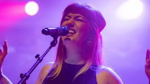 Hayley Sanderson Hayley Sanderson sings into a microphone. She looks passionate and has her arms open and her eyes closed. The background is bathed in purple lighting.