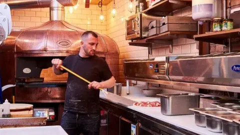 Family handout Man in a dark blue top and jeans standing next to a pizza oven in an Italian restaurant lifting a pizza on to a pizza paddle.
