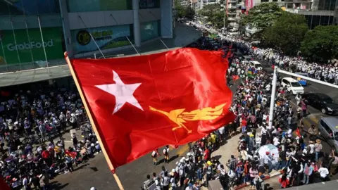 Getty Images The flag of the National League for Democracy party flies over protesters taking part in a demonstration against the February 1 military coup in Yangon