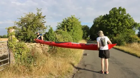 Maddie Rolfe Two men lifting a red kayak over a wall on a country road