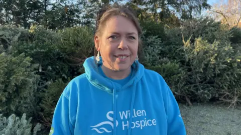Nicola Haseler / BBC A women wearing a blue Willen Hospice sweatshirt smiles at the camera. She has dark hair and wears Christmas tree earrings.