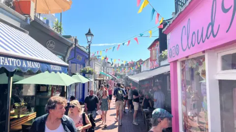A shopping street with dozens of people walking in different directions, colourful bunting strung above the street and small shops painted in different colours.
