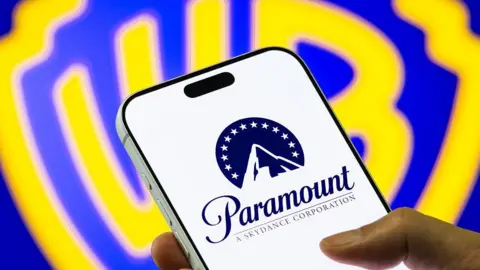 A hand holds a mobile phone, displaying the Paramount logo of a stylised mountain with a halo of stars. In the background is a bright blue and yellow Warner Bros shield logo.