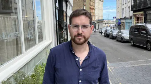 Luke Armitage looking at the camera. He is wearing a blue shirt. The street behind him curves downwards towards a beach.
