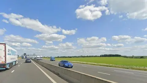 A busy road which has three lanes in each direction. Cars, lorries and vans are being driven along the road. There is a gantry in the distance with road signs that are too far away to read.