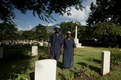 CWGC Shadrack Paull and Lusia Axalte stand holding hands among the graves at the cemetery in Tanzania